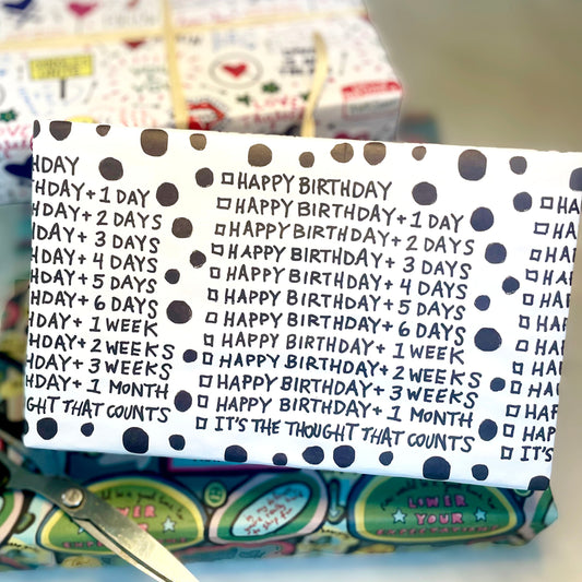 Gift wrap adorned in white paper with black font showing a checklist of different boxes you can check for how late you are in wishing Happy Birthday, like Happy Birthday plus one day, happy birthday plus tow days in handwritten font. The last checked box says, "It's the thought that counts." The background features additional wrapping paper and scissors, creating a scene of festive preparation.