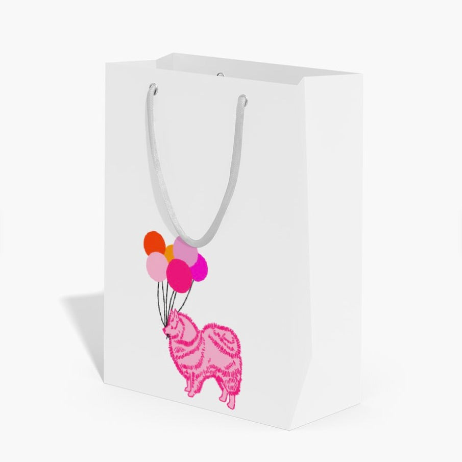 A white paper gift bag with white paper handles, featuring a graphic print of a pink dog holding a cluster of colorful balloons.