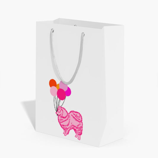A white paper gift bag with white paper handles, featuring a graphic print of a pink dog holding a cluster of colorful balloons.