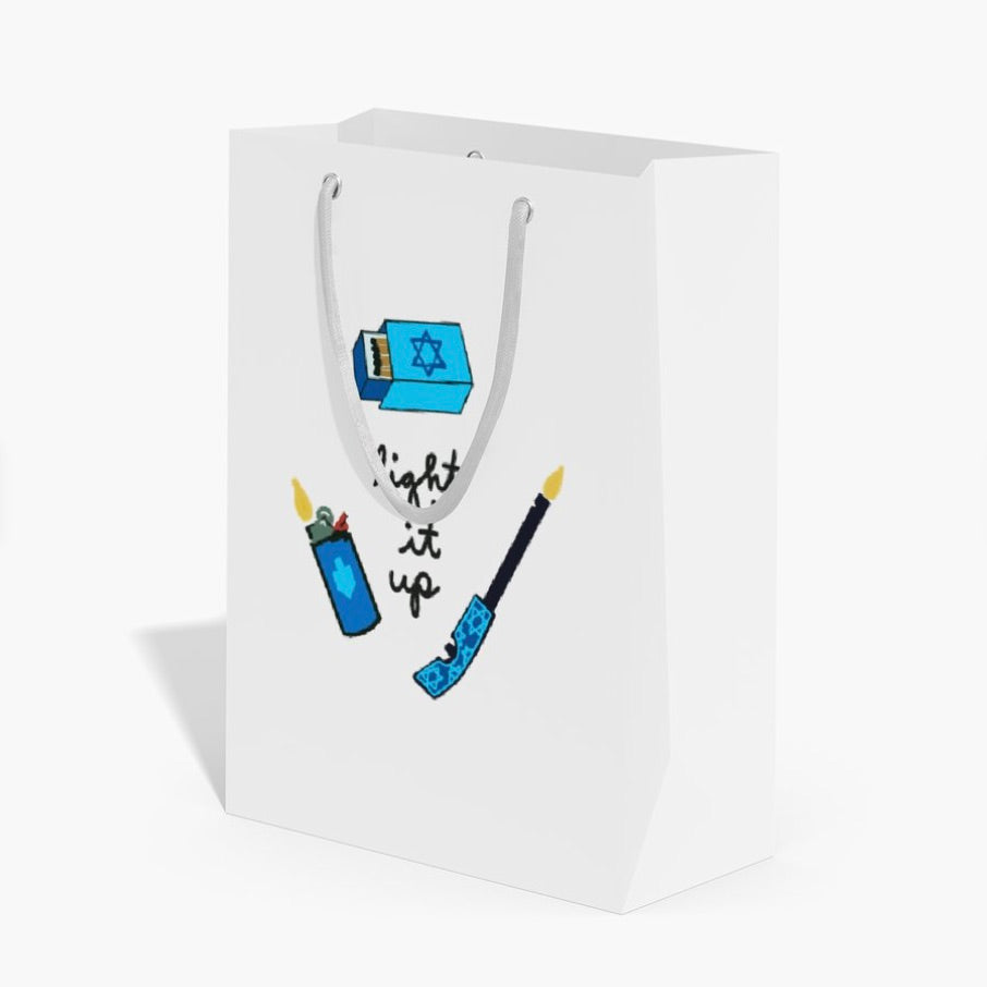 A white paper gift bag with white handles, featuring a printed graphic of blue and black illustrations (a Star of David, a lighter, a candle, and the phrase 'light it up') on a clean white background.
