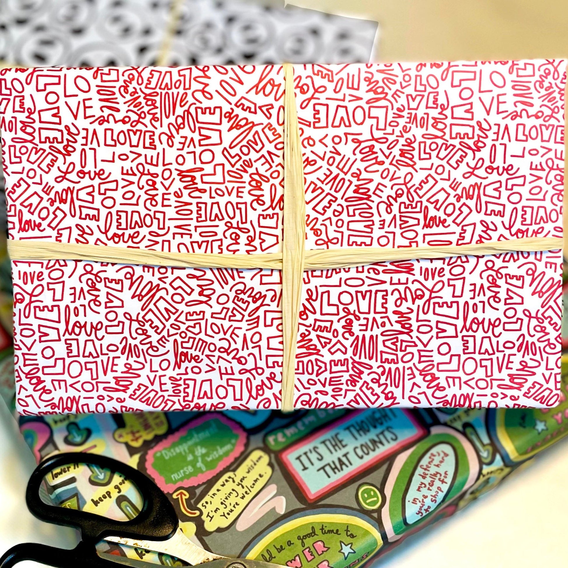 Gift wrap adorned in red and white patterned paper featuring the word "Love" in different handwritten fonts with a cream ribbon. The background features additional wrapping paper and scissors, creating a scene of festive preparation.