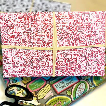Gift wrap adorned in red and white patterned paper featuring the word "Love" in different handwritten fonts with a cream ribbon. The background features additional wrapping paper and scissors, creating a scene of festive preparation.