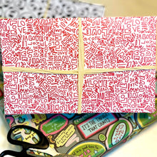 Load image into Gallery viewer, Gift wrap adorned in red and white patterned paper featuring the word &quot;Love&quot; in different handwritten fonts with a cream ribbon. The background features additional wrapping paper and scissors, creating a scene of festive preparation.
